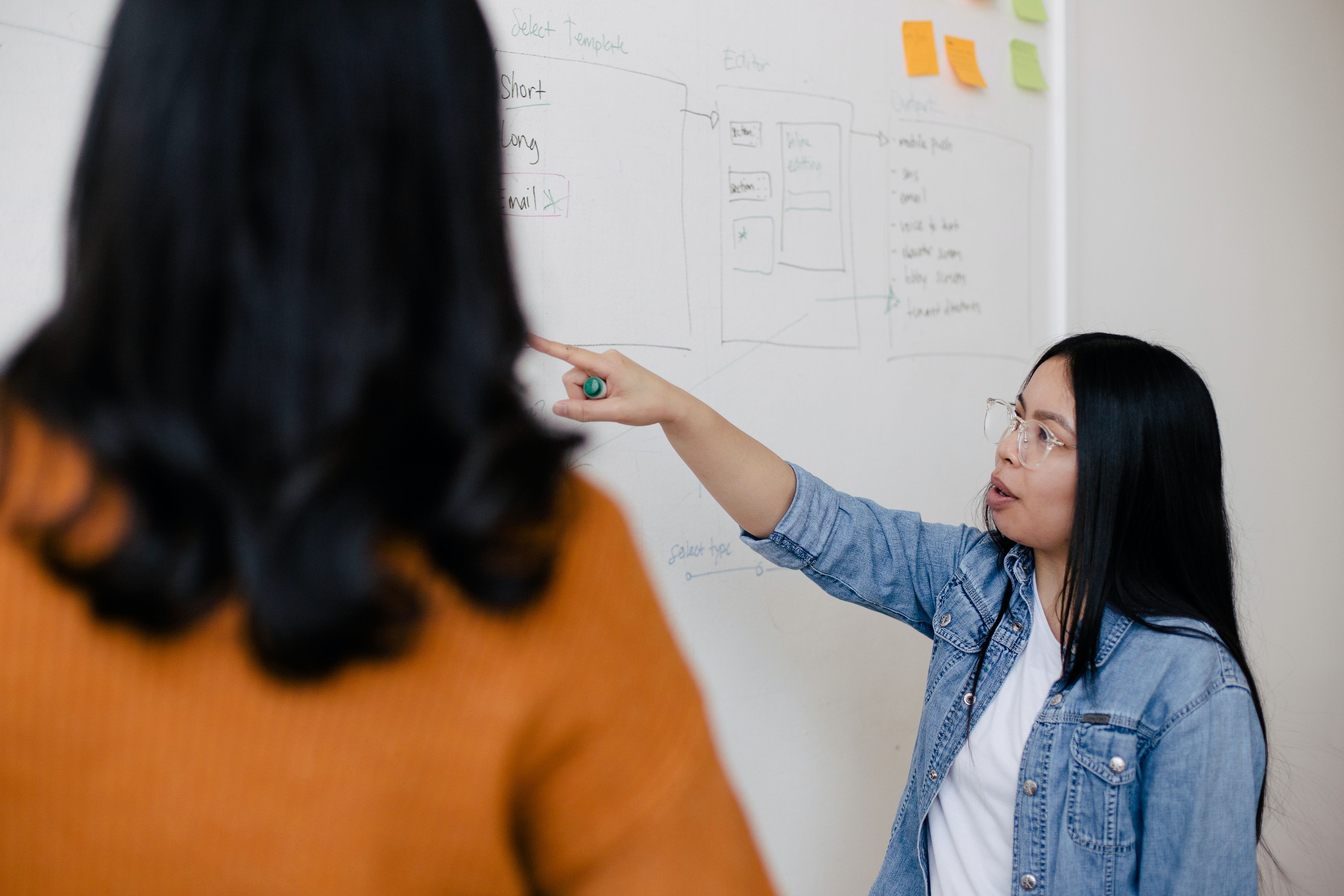 Two women are standing in front of a whiteboard with written notes on it. The woman on the right is pointing something out to the woman on the left.