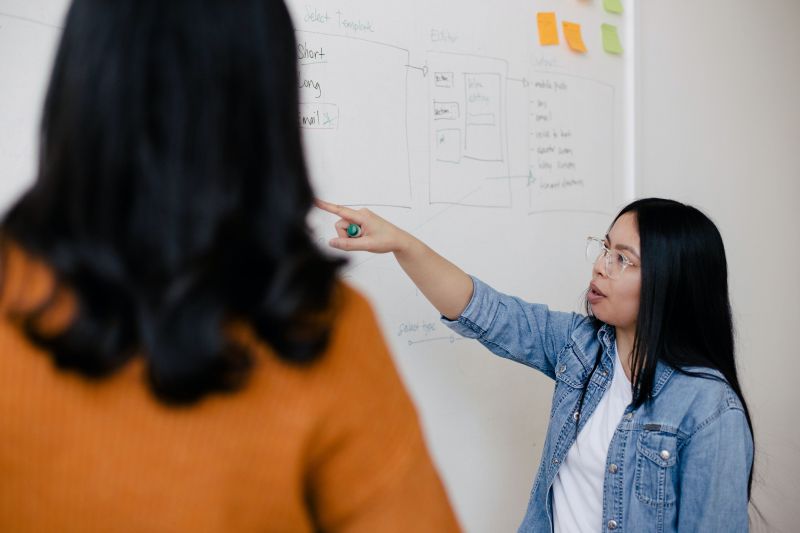 Two women are standing in front of a whiteboard with written notes on it. The woman on the right is pointing something out to the woman on the left.