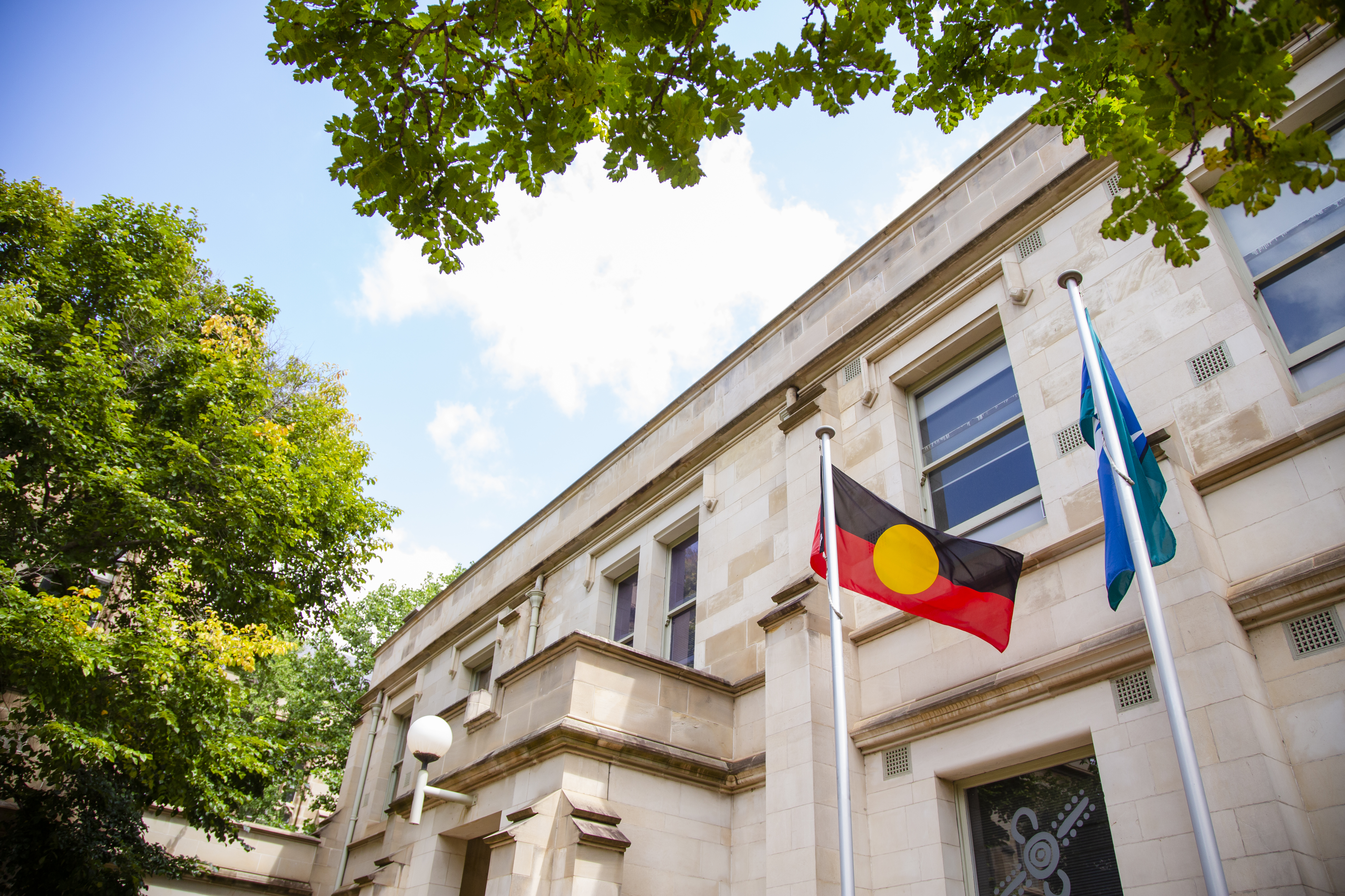 Aboriginal and Torres Strait Islander flags flying on flag poles. There is a sandstone building behind the flags. The photo is taken from close to the ground and pointing upwards at the sky. A blue sky with a couple of clouds is visible and you can see the tops of two trees with green and some yellow leaves.