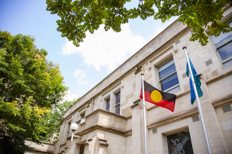Aboriginal and Torres Strait Islander flags flying on flag poles. There is a sandstone building behind the flags. The photo is taken from close to the ground and pointing upwards at the sky. A blue sky with a couple of clouds is visible and you can see the tops of two trees with green and some yellow leaves.